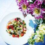 Lättrökt kycklingbröst med sommarsallad på en tallrik. I förgrunden lila och vita blommor.