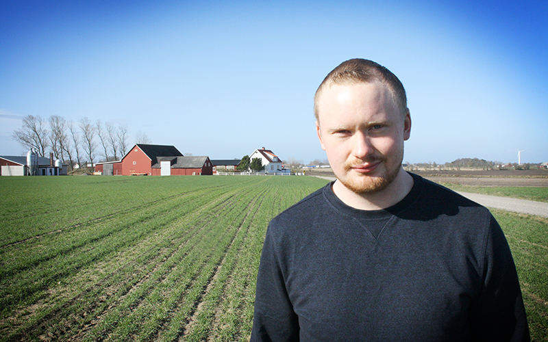 Man ståendes på en åker i svart t-shirt. I bakgrunden syns röda hus.